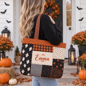 Op de afbeelding: Een gewatteerde tas met een patchworkontwerp in oranje, zwarte en crèmekleurige stoffen met patronen van spoken, pompoenen, spinnenwebben en ruitjes. Het middelste crèmekleurige paneel is geborduurd met de naam Emma in oranje cursief schrift.