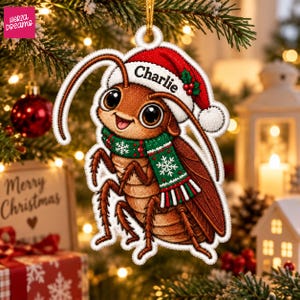 Könnte beinhalten: Ein brauner Kakerlaken-Weihnachtsanhänger, der eine rote Weihnachtsmütze mit dem Namen Charlie auf dem Rand und einen grünen Schal mit weißen Schneeflockenmustern trägt. Der Anhänger hat einen weißen gestickten Rand und eine goldene Aufhängeschnur.