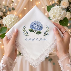 May include: A white square handkerchief with a lace border, featuring an embroidered blue hydrangea flower flanked by two green leafy sprigs. Below the floral design, the names Kayleigh & Audra are embroidered in blue script lettering.