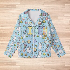Peut inclure: Haut de pyjama bleu clair sur le th&egrave;me de P&acirc;ques avec une bordure blanche. Le haut de pyjama pr&eacute;sente un motif r&eacute;p&eacute;t&eacute; de lapins blancs, d'&oelig;ufs d&eacute;cor&eacute;s et de papillons. Le nom "AIDEN" est imprim&eacute; sur la poitrine en lettres color&eacute;es. Le pyjama a des manches longues et un col.