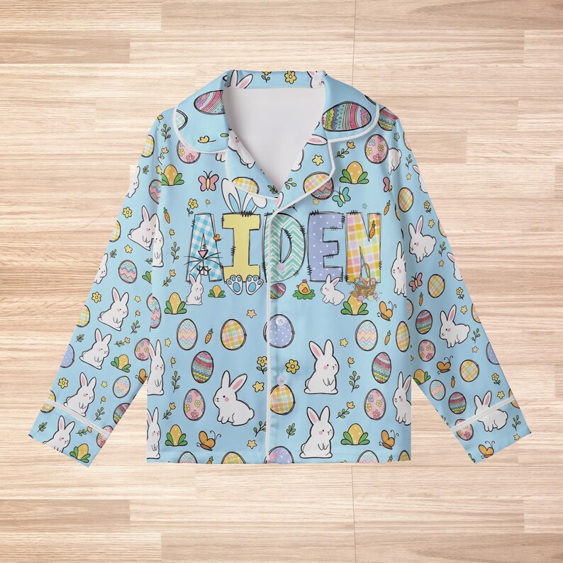 Peut inclure: Haut de pyjama bleu clair sur le th&egrave;me de P&acirc;ques avec une bordure blanche. Le haut de pyjama pr&eacute;sente un motif r&eacute;p&eacute;t&eacute; de lapins blancs, d'&oelig;ufs d&eacute;cor&eacute;s et de papillons. Le nom "AIDEN" est imprim&eacute; sur la poitrine en lettres color&eacute;es. Le pyjama a des manches longues et un col.