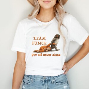Puede incluir: Camiseta blanca con el texto "TEAM PUNCH you are never alone" en marrón. El gráfico muestra un mono gris golpeando un peluche marrón. La camiseta está hecha de un material suave.
