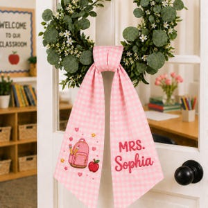 Könnte beinhalten: Ein rosa-weiß kariertes Stoffband, das an einem grünen Kranz mit weißen Blumen hängt. Das Band zeigt gestickte Motive eines rosa Rucksacks, Bleistiften und eines roten Apfels sowie den in rosa Garn gestickten Text MRS. Sophia.
