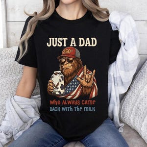 Op de afbeelding: Zwart t-shirt met een afbeelding van Bigfoot met een zonnebril en een baseballpet, met een melkpak en een Amerikaanse vlag. Tekst luidt "JUST A DAD" en "Who Always Came Back With the Milk."