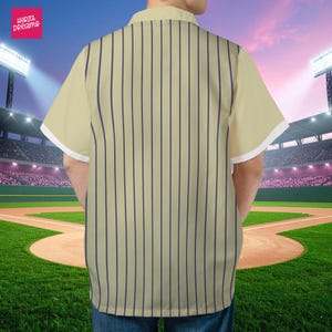 K&ouml;nnte beinhalten: Cremefarbenes Hemd mit vertikalen marineblauen Streifen. Das kurz&auml;rmelige Hemd hat wei&szlig;e Verzierungen an den &Auml;rmeln. Das Hemd ist von "Herza Dreams". Der Hintergrund ist ein Baseballfeld.