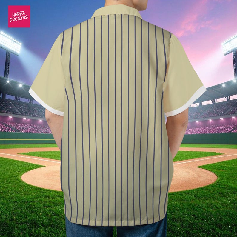 K&ouml;nnte beinhalten: Cremefarbenes Hemd mit vertikalen marineblauen Streifen. Das kurz&auml;rmelige Hemd hat wei&szlig;e Verzierungen an den &Auml;rmeln. Das Hemd ist von "Herza Dreams". Der Hintergrund ist ein Baseballfeld.