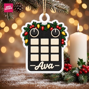 May include: A rectangular Christmas ornament featuring a black grid pattern with nine cream-colored squares, two black circular knobs at the top, and a festive garland of green leaves, red berries, and colorful light bulbs. The name Ava is printed in white.