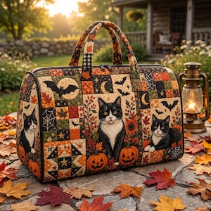 Op de afbeelding: Een gewatteerde reistas met een patchworkpatroon van zwart-witte katten, oranje pompoenen, herfstbladeren, vleermuizen en maansikkels. De tas heeft twee handvatten, een ritssluiting en een zwarte bies, met een seizoensgebonden herfstthema.