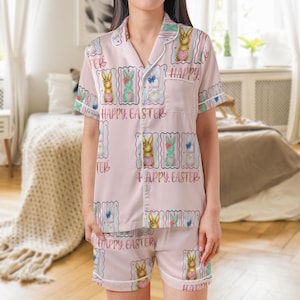 K&ouml;nnte beinhalten: Hellrosa Pyjama-Set mit einem kurz&auml;rmeligen Hemd und Shorts. Das Set zeigt ein sich wiederholendes Muster von Osterhasen, die Herzen und Schmetterlinge halten, sowie die Worte "Happy Easter". Wei&szlig;e Paspeln akzentuieren den Kragen, die Manschetten und die Taschen.