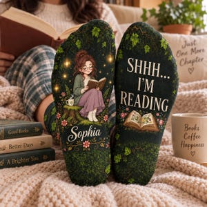 May include: A pair of dark green socks featuring a printed illustration of a person reading a book on a tree stump, surrounded by ivy, flowers, and glowing lanterns. The name Sophia is printed below, and the text SHHH... I'M READING appears above an open book.