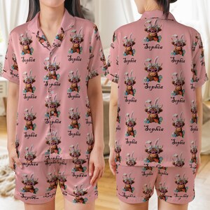 May include: Pink pajama set featuring a repeating pattern of a cartoon Highland cow wearing sunglasses and bunny ears, with the name "Sophia" in cursive. The set includes a button-down shirt and shorts.