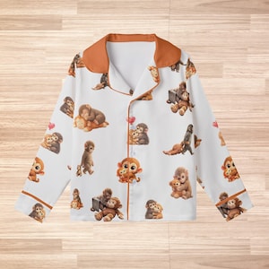 May include: White pajama top with a repeating pattern of cartoon monkeys in various poses. The long-sleeved top has a brown collar and trim. The monkeys are in shades of brown and orange, with some holding hearts.