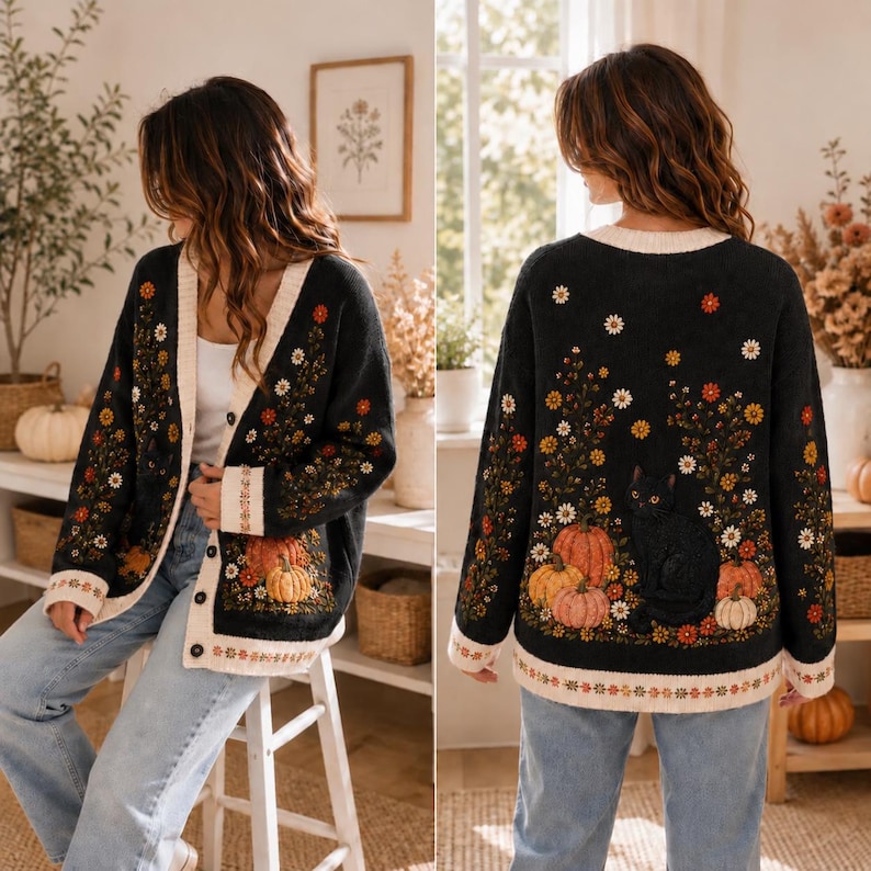 K&ouml;nnte beinhalten: Eine schwarze Strickjacke mit Kn&ouml;pfen und einem Strickmuster aus orangefarbenen K&uuml;rbissen, einer schwarzen Katze und bunten Wildblumen. Das Kleidungsst&uuml;ck hat einen cremefarbenen Saum am V-Ausschnitt, an den &Auml;rmelb&uuml;ndchen und am unteren Rand.