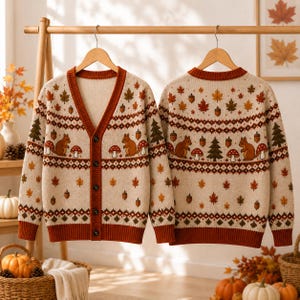 May include: A beige V-neck cardigan featuring a repeating autumn-themed pattern of brown squirrels, red and white mushrooms, green pine trees, acorns, and orange maple leaves. The garment has a dark orange ribbed trim, cuffs, and buttons.