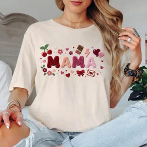 May include: Cream-colored t-shirt with the word "MAMA" in textured, plush letters. The design includes cherries, flowers, hearts, a chocolate bar, and a bow. The shirt is a casual, everyday wear item.