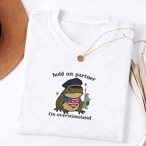 Puede incluir: Camiseta blanca con una rana de dibujos animados que lleva un gorro azul marino y un pañuelo con la bandera estadounidense, sosteniendo la Estatua de la Libertad. El texto "hold on partner, I'm overstimulated" está bordado en la camiseta. Un collar dorado está drapeado.