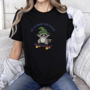 Op de afbeelding: Zwart t-shirt met een wasbeer op een skateboard, met een groene hoed. De tekst "OFF TO COMMIT DAD FOOLERY" staat boven de wasbeer. Het skateboard is rood en geel.