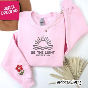 May include: A light pink sweatshirt with the text "BE THE LIGHT" and "MATHEW 5:14" embroidered in a dark color. The design includes a sun and waves. A small flower is embroidered on the sleeve. The word "embroidery" is written on the bottom right.
