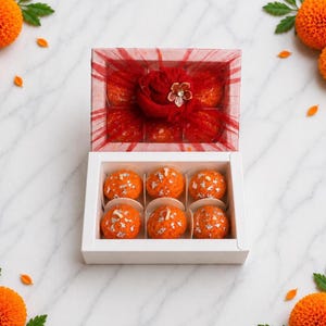 May include: A white rectangular gift box containing six round orange sweets topped with silver leaf flakes. The lid is transparent with a red mesh fabric overlay, featuring a large red fabric flower decoration with a central metallic accent.
