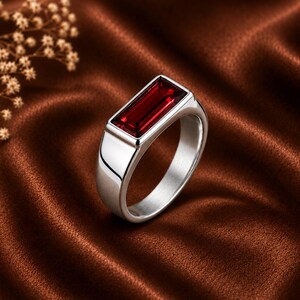 May include: A silver-toned ring featuring a rectangular, deep red gemstone set horizontally in a smooth, polished metal band. The design showcases a clean, geometric aesthetic with a prominent, faceted stone that reflects light across its surface.