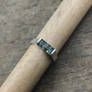 May include: A silver ring with a rectangular, moss-green gemstone. The ring is set on a wooden dowel, showcasing its design. The gemstone has a natural, organic pattern, and the ring has a simple, modern band.