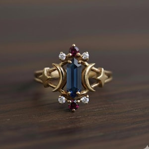 Peut inclure: Bague dorée ornée d'une pierre centrale rectangulaire bleu foncé. La bague est ornée de petites pierres rouges et blanches, de motifs de croissant de lune et d'étoiles. La bague est posée sur une surface en bois sombre.