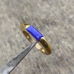 May include: A gold ring with a rectangular, deep blue stone set in the center. The ring has a smooth, polished finish and is held by a metal tool against a neutral background. The ring's design is simple yet elegant.