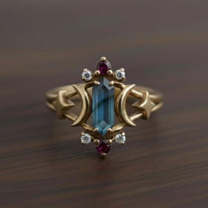 May include: A gold-colored ring featuring a central, rectangular, teal-colored gemstone. The ring is adorned with small, round, white and red gemstones, and crescent moon and star designs. The ring is set against a dark brown wooden surface.