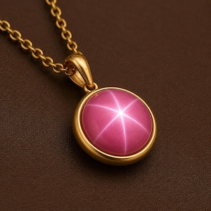 May include: A gold pendant necklace featuring a round, cabochon-cut pink gemstone with a prominent six-rayed star effect centred on the stone. The pendant is held by a polished gold bail and hangs from a matching gold link chain.