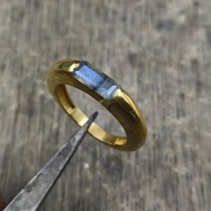May include: A gold ring with a rectangular, blue-gray gemstone. The ring is held by a pair of metal tweezers, showcasing the craftsmanship. The band has a smooth, polished finish.