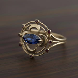 May include: A gold-colored ring featuring a large, oval-shaped blue gemstone at its center. The ring is adorned with a circular design incorporating crescent moon shapes and small red gemstones. The ring's band is a simple, polished gold.