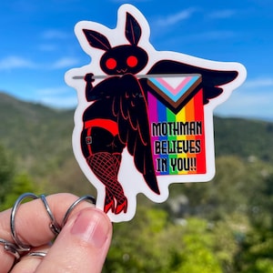 May include: A sticker featuring a black moth-like creature with red eyes and a red garter belt, holding a flag with the Pride Progress flag design. The flag reads "Mothman Believes In You!!"