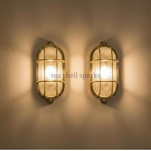 Op de afbeelding: Twee messingkleurige, ovaalvormige wandkandelaars met een rasterpatroon over het glas. De wandkandelaars zijn verlicht en werpen een warme gloed op de muur. De tekst "sea shell sparks" is zichtbaar.
