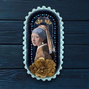 May include: A decorative oval-shaped wall art piece featuring a print of Johannes Vermeer's "Girl with a Pearl Earring." The artwork is set within a dark blue frame, adorned with light blue pom-poms and gold-colored roses at the bottom.