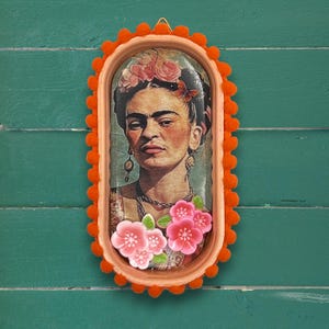May include: An oval-shaped wall art piece featuring a portrait of Frida Kahlo, framed in orange with a pom-pom trim. The artwork includes pink flowers and a butterfly, set against a green background. The piece is ready to hang.
