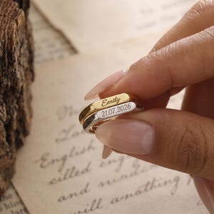 May include: Two stacked metal rings, one gold-toned and one silver-toned, featuring flat rectangular surfaces. The gold ring is engraved with the name Emily, and the silver ring is engraved with the date 21.07.2026 in a clear, sans-serif font.