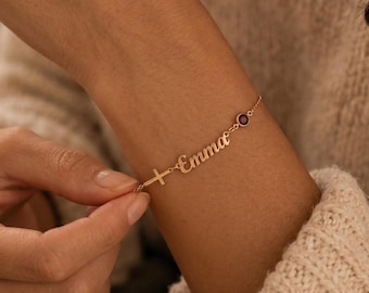 Puede incluir: Una pulsera de cadena dorada que presenta un pequeño dije de cruz, el nombre Emma en letra cursiva y una piedra preciosa redonda de color rojo oscuro. Los delicados eslabones metálicos conectan estos tres elementos en línea.