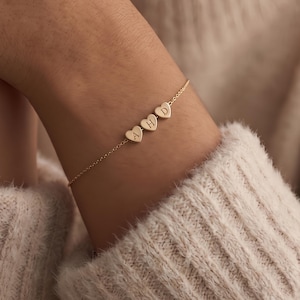 Puede incluir: Una pulsera dorada con tres colgantes en forma de corazón, cada uno con una letra. La pulsera, con las letras "AHD", se lleva en la muñeca. Una joya delicada y elegante.