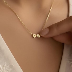 Puede incluir: Un delicado collar de oro con dos pequeños colgantes en forma de corazón. El collar se lleva puesto, con la cadena sobre la piel. Los colgantes están juntos, creando un diseño sutil y elegante. Una joya delicada.