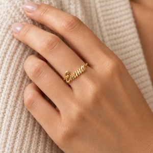 Può includere: Un anello color oro che presenta il nome Emma in un carattere corsivo. L'anello ha una fascia sottile e lucida che si collega alla scritta, posizionata al centro del dito. Il metallo ha una finitura liscia e riflettente.