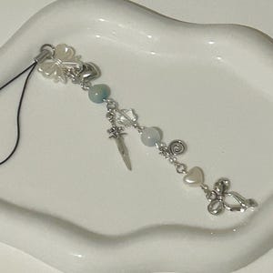 Puede incluir: Un delicado dije para teléfono con dijes plateados de lazo, corazón y flores, junto con cuentas azul claro y un pequeño colgante de espada. El dije está sujeto a un lazo de cordón negro.