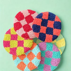 May include: A collection of colorful, round coasters with a checkerboard pattern. The coasters are in shades of red, pink, blue, orange, yellow, green, and purple. They are made of a textured material and are arranged on a light green background.
