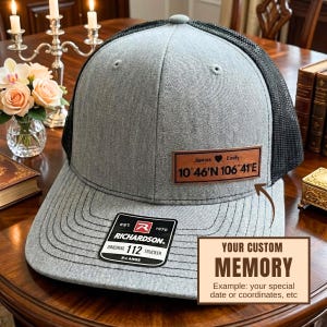 May include: A gray trucker hat with a black mesh back and a curved brim. The hat features a leather patch with the names "James" and "Emily" and coordinates. The hat also has a "Richardson" tag. The text "YOUR CUSTOM MEMORY" is also visible.