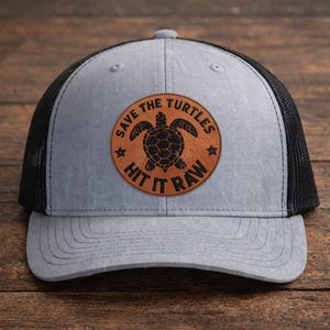 May include: A gray trucker hat with a black mesh back. The front features a circular brown leather patch with a black sea turtle design and the text "SAVE THE TURTLES HIT IT RAW".