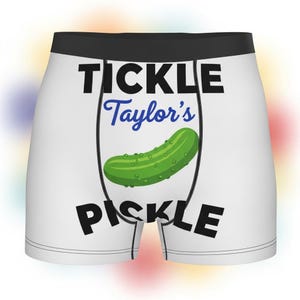 May include: White boxer briefs with a black waistband. The front features the text "TICKLE Taylor's PICKLE" in black and blue, with a green pickle graphic in the center. The background is blurred with colorful spots.