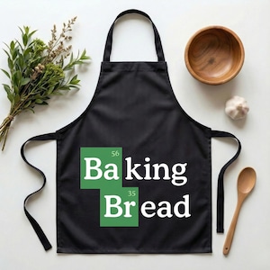 Op de afbeelding: Zwarte schort met de witte tekst "Baking Bread", gestileerd als een periodiek systeem. Een houten kom, lepel, knoflook en groen omringen de schort.