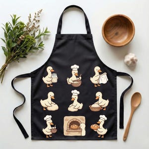 May include: Black apron featuring cartoon ducks in chef hats, baking bread. The ducks are depicted in various baking activities, such as mixing ingredients, kneading dough, and removing bread from an oven. A wooden bowl, spoon, and garlic are also visible.