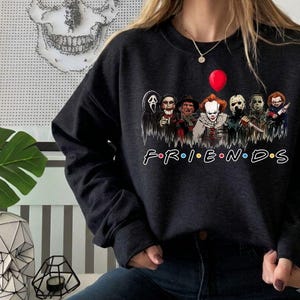 May include: A black crewneck sweatshirt featuring a graphic print of various horror movie characters standing together under a red balloon. Below the characters, the word FRIENDS is printed in a white font with colorful dots between each letter.