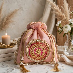 May include: A light pink drawstring pouch featuring a central circular medallion with magenta fabric, gold embroidery, and reflective mirror accents. The bag is trimmed with gold metallic lace, pearl-like beads, and two gold tassels on drawstrings.