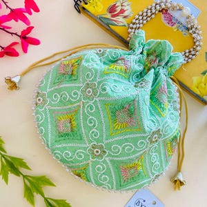 May include: A round, mint green drawstring pouch with detailed embroidery in white, yellow, pink, and gold. The pouch has a beaded edge and gold-coloured drawstring cords. A small tag with the text "BYRA" is attached.